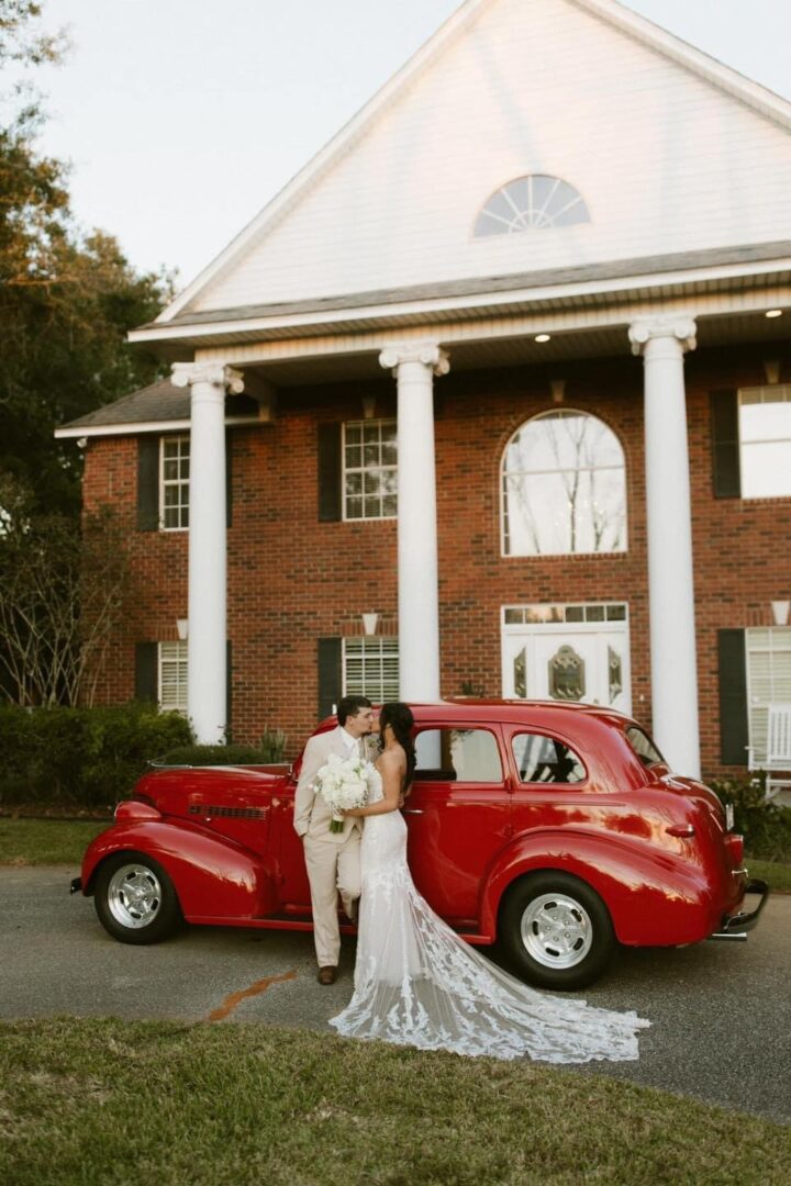 Bride and groom kissing by red car.